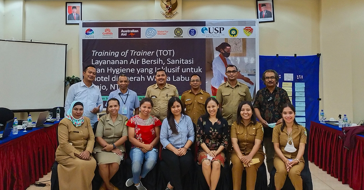 A group is assembled indoors with a row standing and a row sitting, they are participants in the local stakeholder training on GEDSI WASH in Labuan Bajo