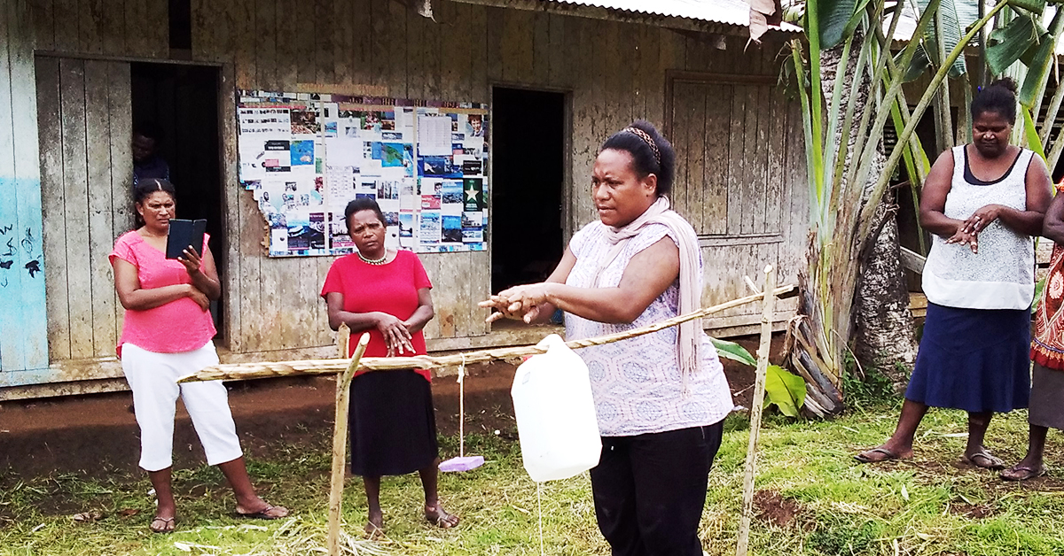 A woman in PNG is demonstrating how to wash your hands properly using a tippy tap device and people watch on