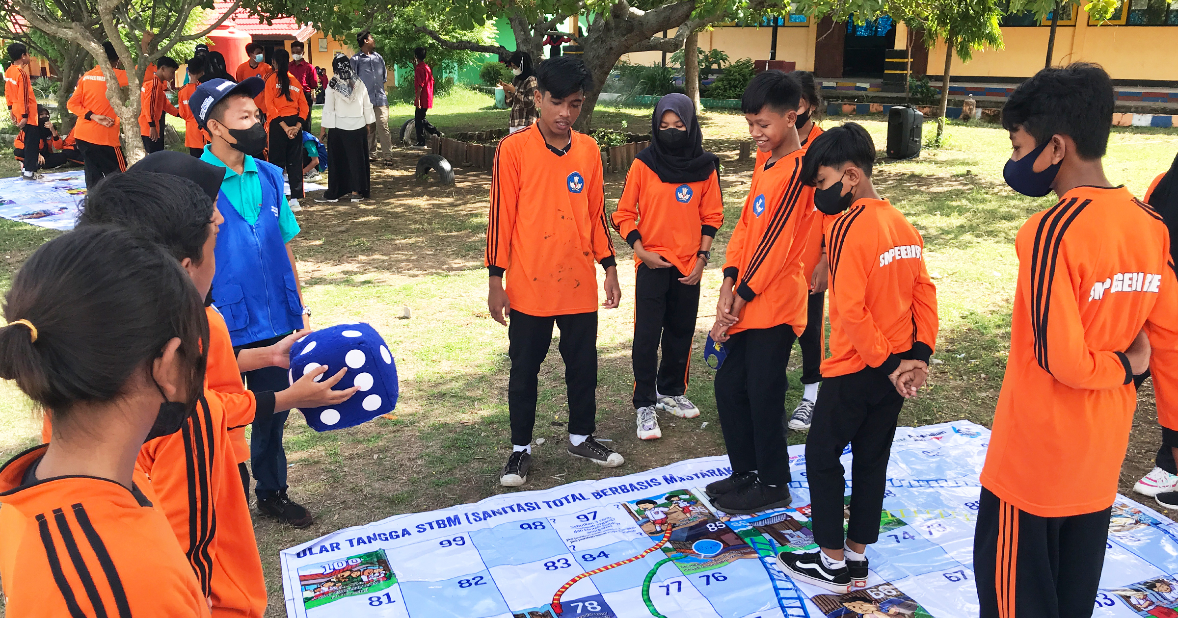 A young school boy in a blue vest stands with other children in orange and black uniforms. They are outside at their school. One child holds a giant blue dice and they all stand on a large mat on the ground which is a big game of snakes and ladders – used to help shifting stigma and taboos around MHH