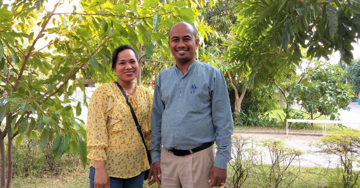 A Cambodian man and woman smile at the camera with greenery behind them, they are husband and wife